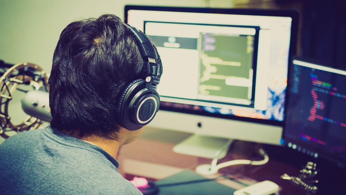Développeur de dos, casque sur les oreilles, devant un iMac et un second écran de code